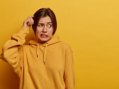 Unsure doubtful woman scratches head, has amnesia, makes tough decision, looks troubled aside, stands puzzled, clenches teeth, dressed in hoodie, poses against yellow wall, empty space for text